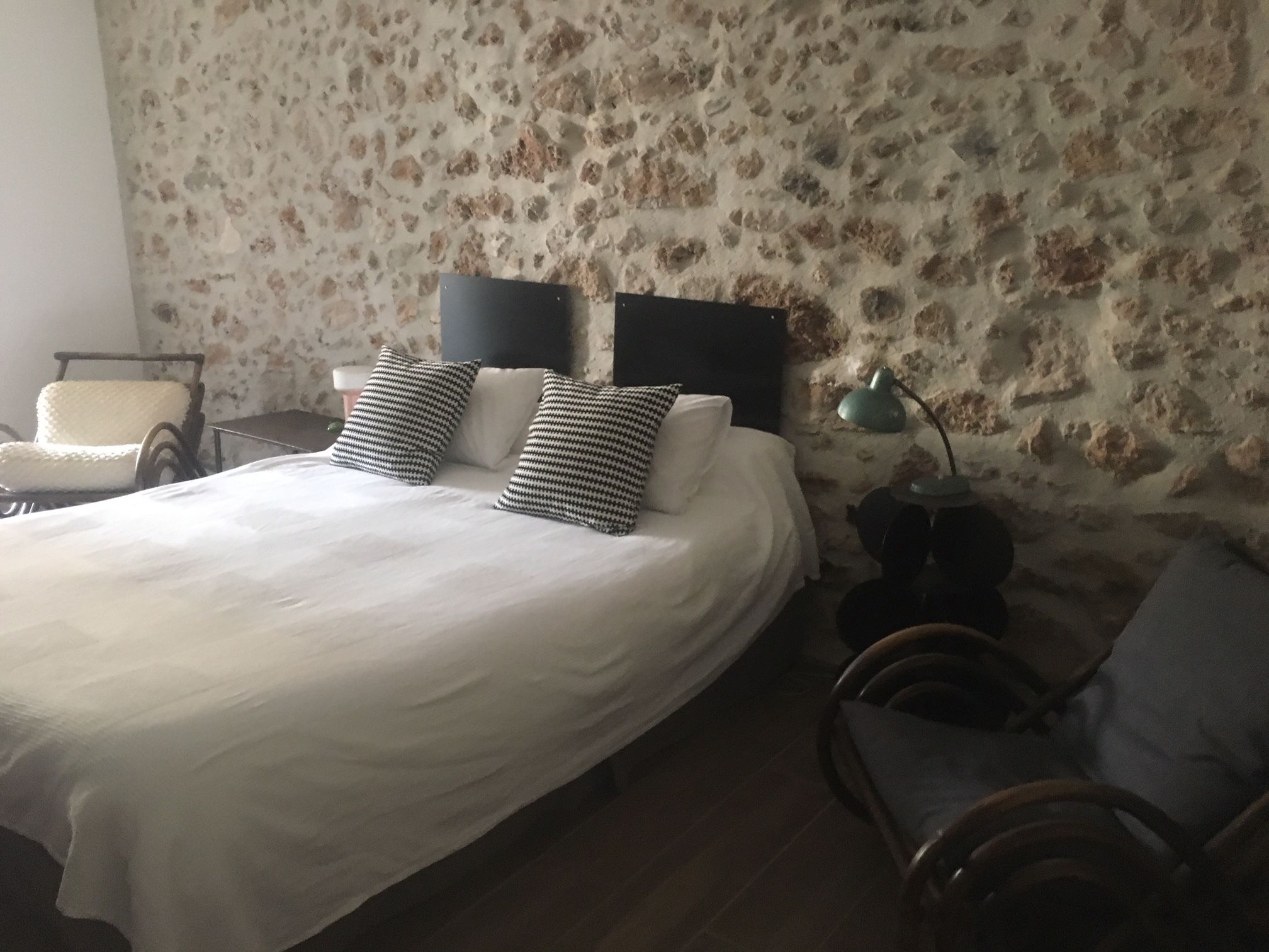 A bedroom with a stone wall, a bed with white bedding, two black headboards, a chair, a small table, and a lamp.