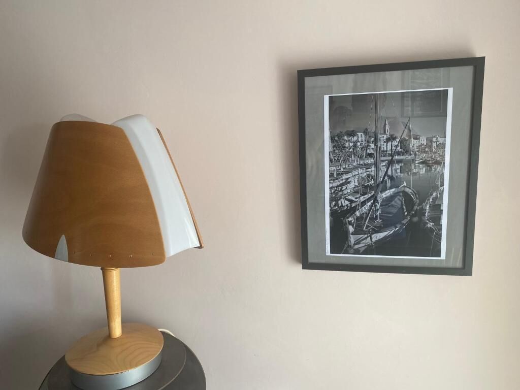 A lamp with a wooden shade and a framed black and white photograph of a harbor scene on the wall.
