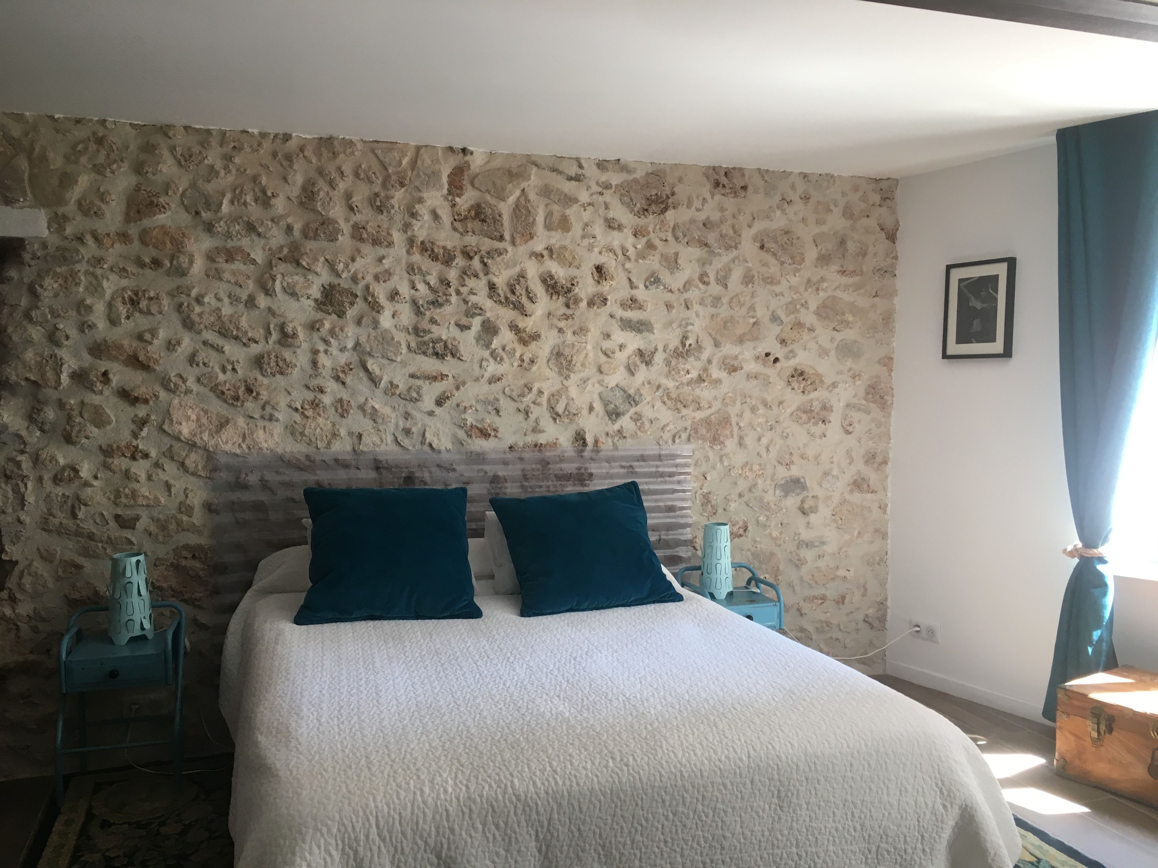 Un dormitorio con pared de piedra, una cama con ropa de cama blanca y dos almohadas turquesa, una mesita de noche con una lámpara y un aparador de madera.