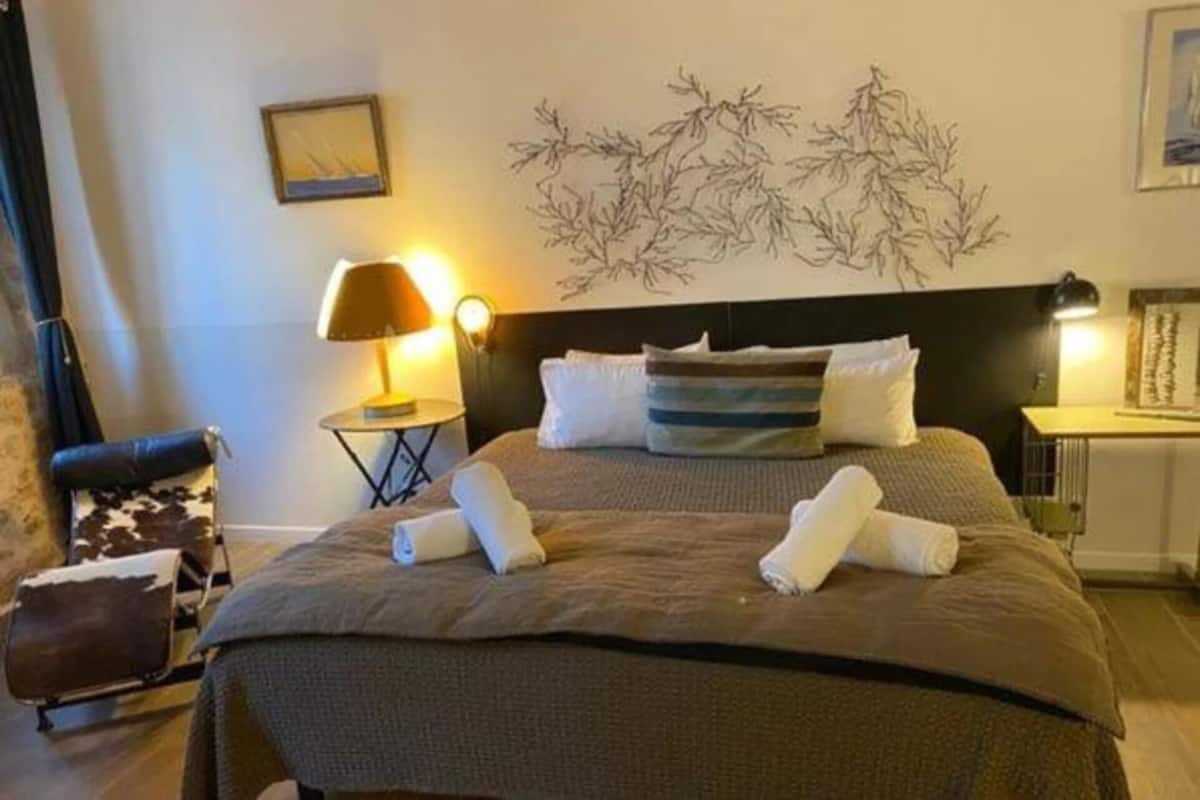 Ein ordentlich bezogenes Bett mit einer dunkelbraunen Tagesdecke und zwei aufgerollten Handtüchern darauf. Ein kleiner runder Tisch mit einer Lampe steht daneben.