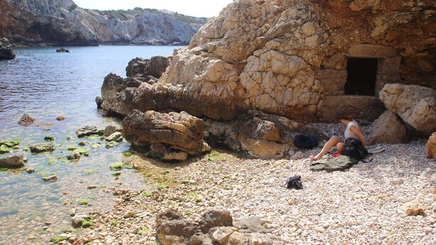 A rocky beach with clear water, a cave-like structure, and a person sitting on the shore.