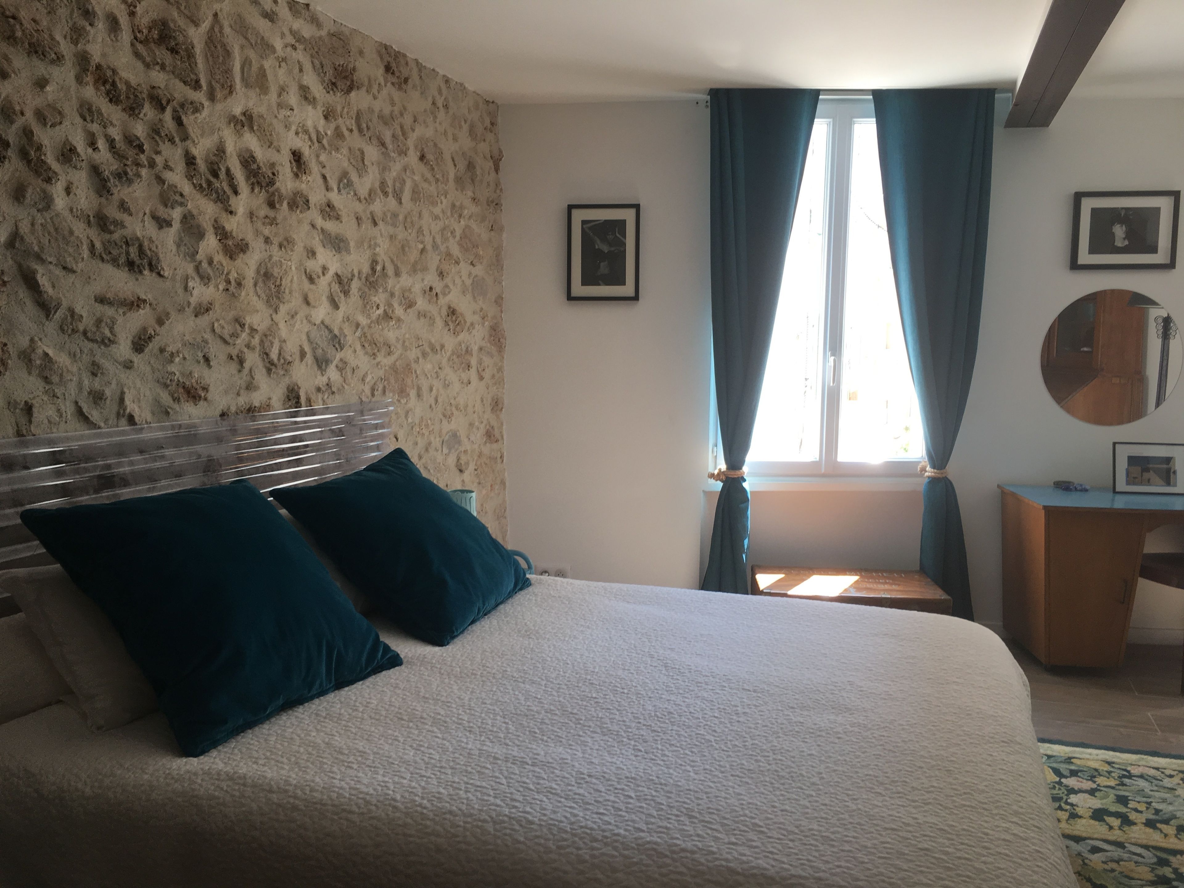 A bedroom with a stone wall, a bed with two pillows, a desk, and a window with curtains.