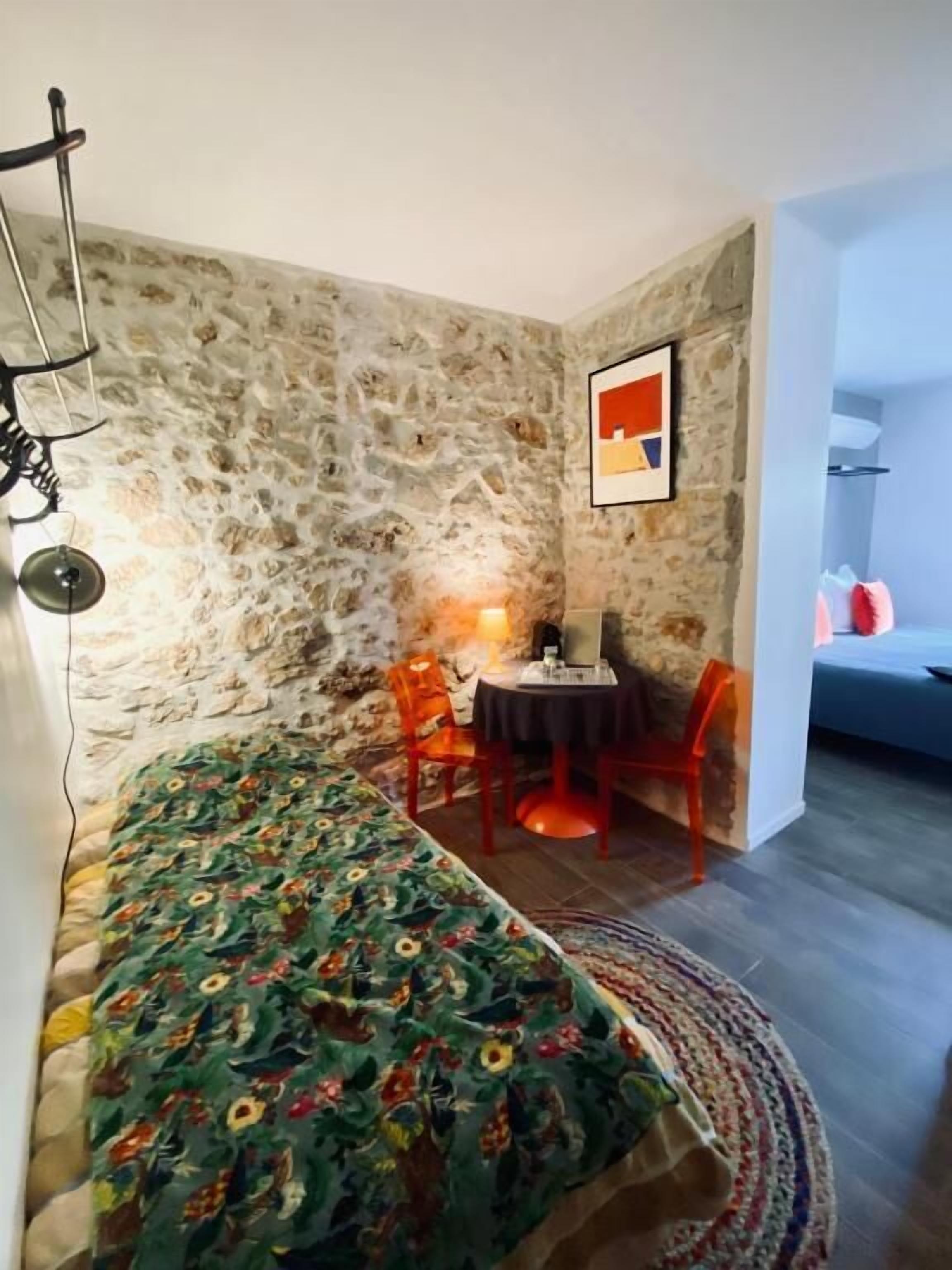 A cozy room with a stone wall, a bed with a floral bedspread, a small table with a lamp, and a chair.