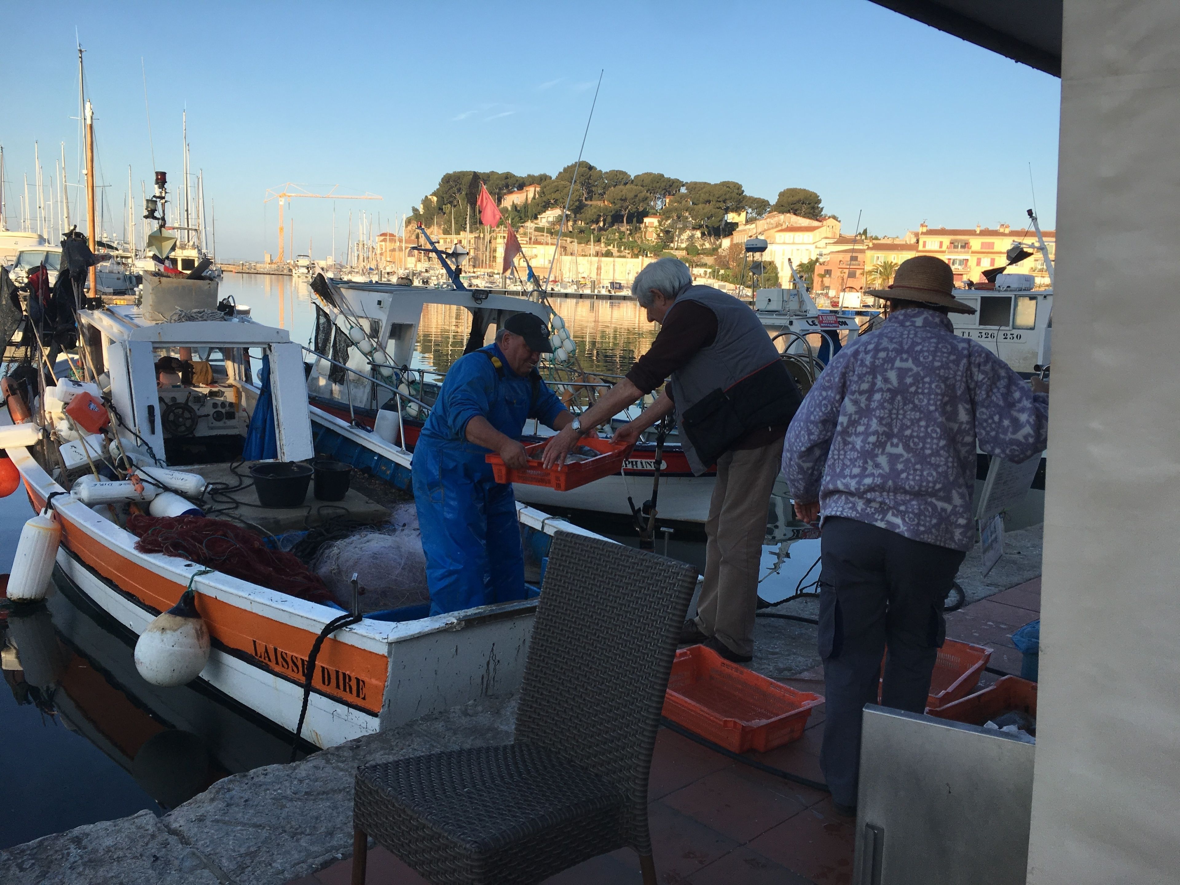 Una marina con barche ormeggiate, persone al lavoro e edifici sullo sfondo.