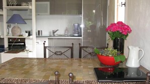 Un coin repas avec une table à carreaux, des chaises, un vase de fleurs, une carafe, et une cuisine en arrière-plan.