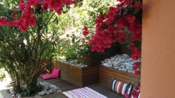Une terrasse en bois avec un espace salon, entourée d’une végétation luxuriante et de fleurs rouges.