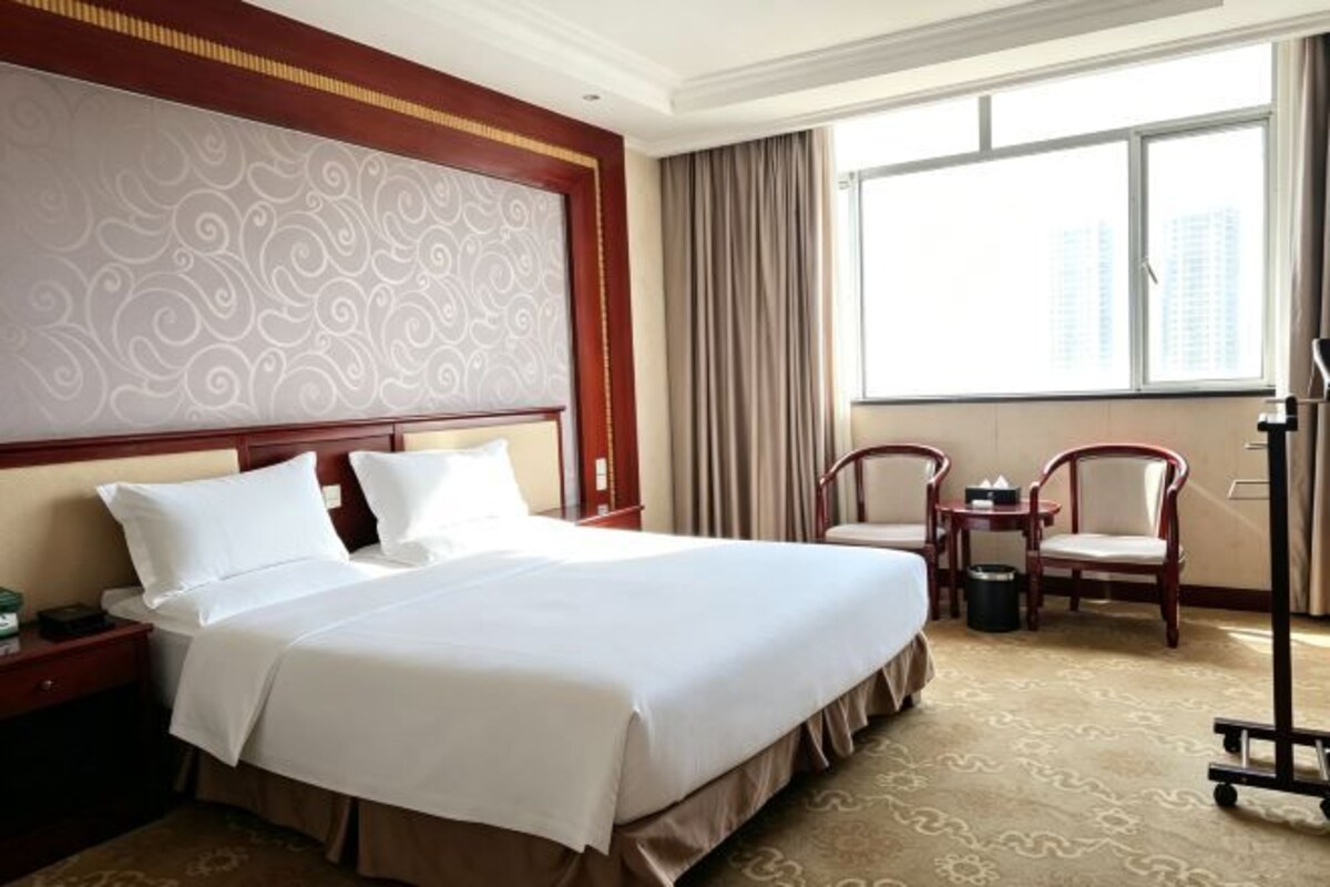 Habitación de hotel con una cama grande, un escritorio, dos sillas, un ventanal con cortinas y vistas a edificios.