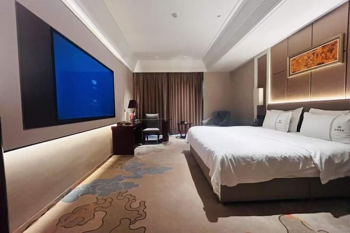 Una habitación de hotel con un televisor de pantalla plana grande, una cama con ropa blanca, un escritorio con lámpara y una silla.