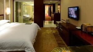 A hotel room with a bed, a television, a desk, and a glass table with a fruit bowl. - West Tour Hotel (Shanshan)