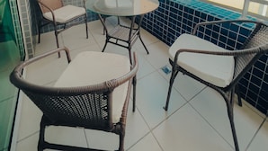 Een balkon met rieten stoelen en een glazen tafel, met uitzicht op een strand en een hoog gebouw.