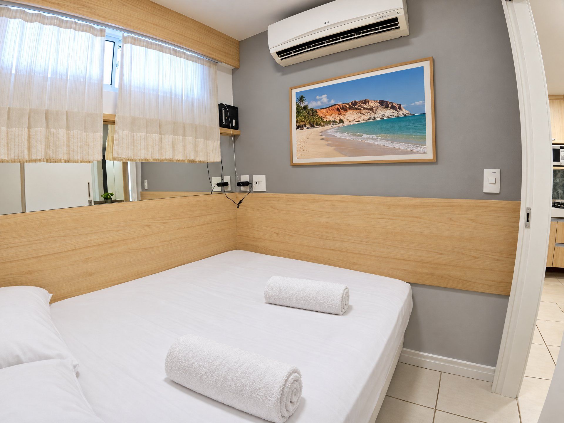 A hotel room with a bed, an air conditioner, a painting of a beach, and a view of the kitchen.