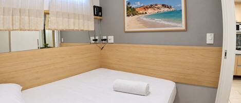 A hotel room with a bed, an air conditioner, a painting of a beach, and a view of the kitchen.