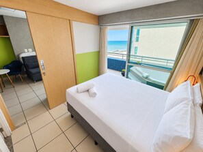 A hotel room with a bed, a chair, a desk, and a view of the sea.