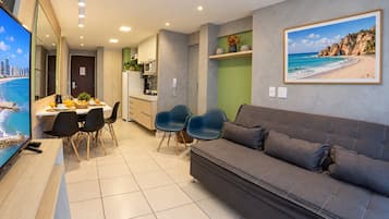 Una sala moderna con un sofá gris, un comedor con mesa y sillas, una cocina con armarios y un televisor grande mostrando una escena de playa.