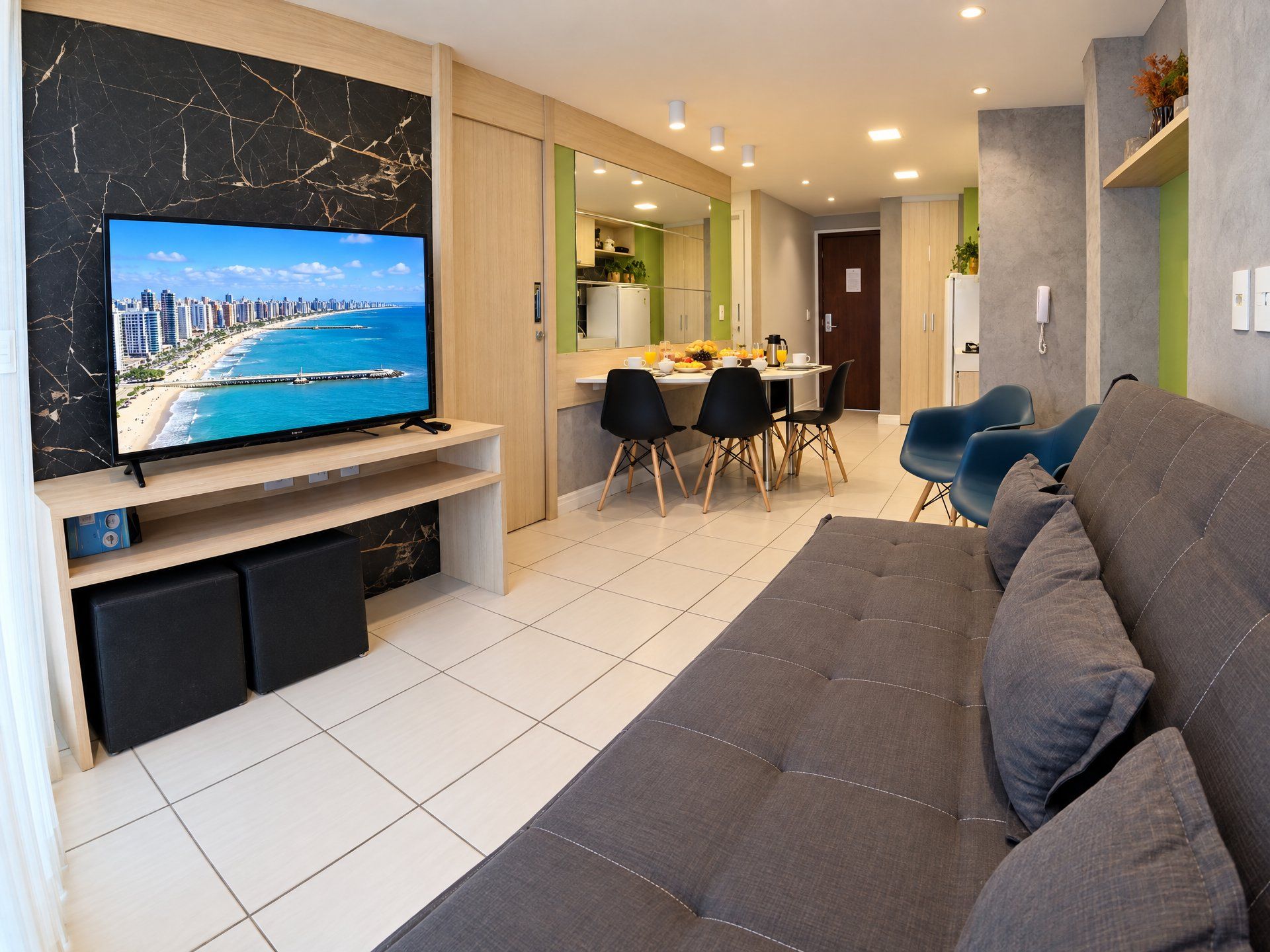 A modern living room with a flat-screen TV, a sofa, a dining area with chairs, and a kitchen in the background.