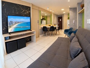 A modern living room with a flat-screen TV, a sofa, a dining area with chairs, and a kitchen in the background.