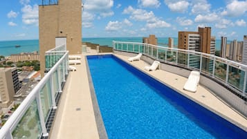Piscina no terraço com água azul cristalina, rodeada por um guarda-corpo de vidro e vista para um horizonte urbano com edifícios e o mar.