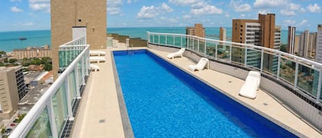 Una piscina en la azotea con agua cristalina, rodeada por una barandilla de vidrio, con vistas a un paisaje urbano que incluye edificios y el mar.