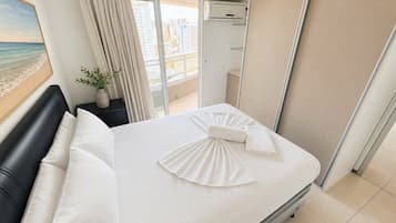 Quarto de hotel com cama, mesa de cabeceira, planta em vaso e vista da cidade pela janela.