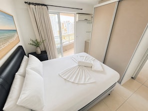 A hotel room with a bed, a nightstand, a potted plant, and a view of the city through the window. - Apartamento Vista mar Praia de Iracema (Fortaleza)