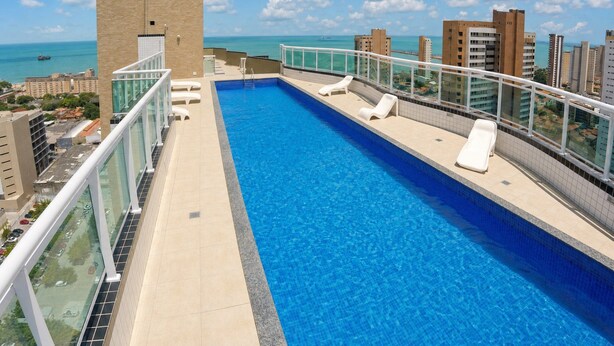 Ein Pool auf dem Dach mit klarem, blauem Wasser, umgeben von einem Glasgeländer und mit Blick auf eine Stadtlandschaft mit Gebäuden und dem Meer.