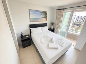 A hotel room with a bed, a nightstand, a painting of a beach, a potted plant, and a view of buildings through a window. - Apartamento Vista mar Praia de Iracema (Fortaleza)