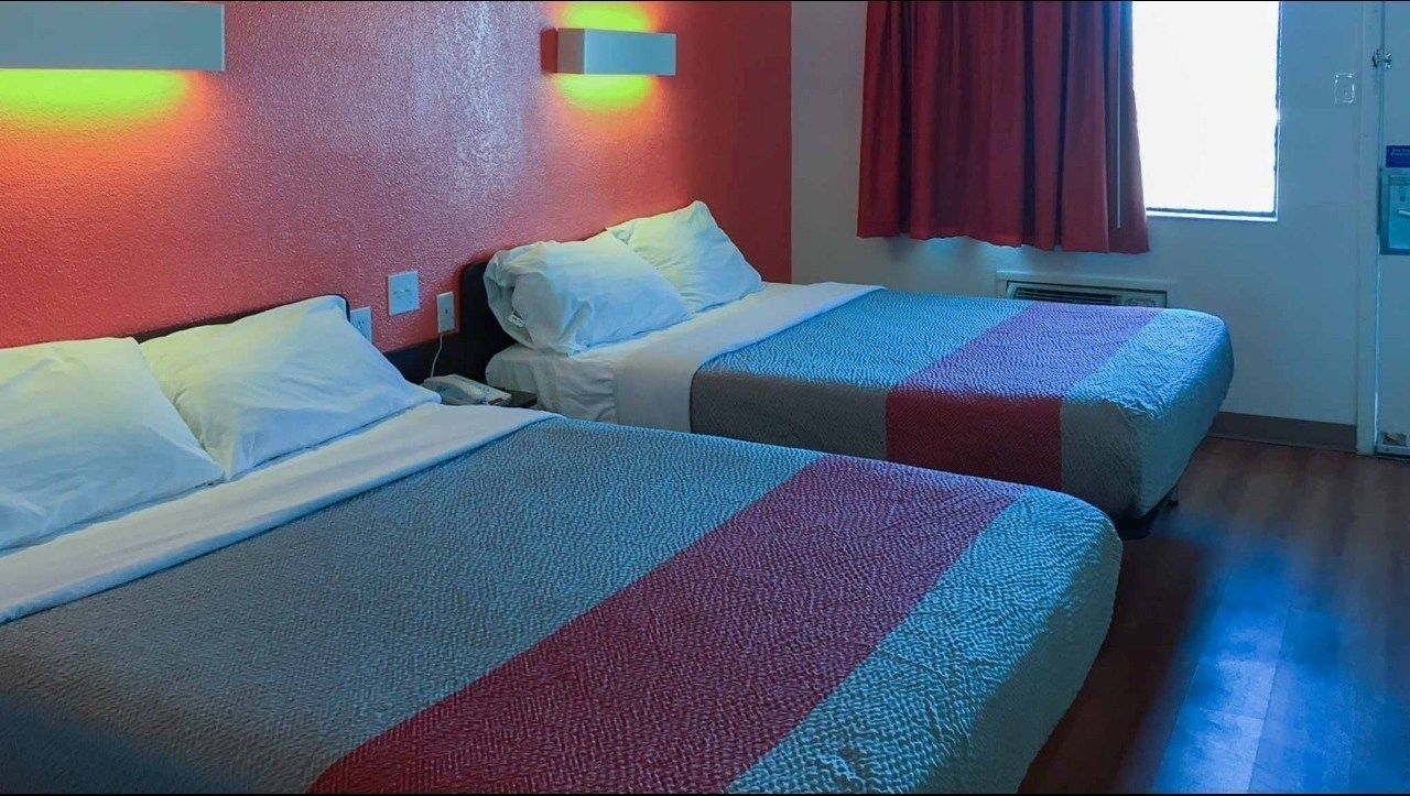A hotel room with two beds, each with white linens and a colorful striped blanket. There is a window with red curtains and a wall-mounted light fixture.