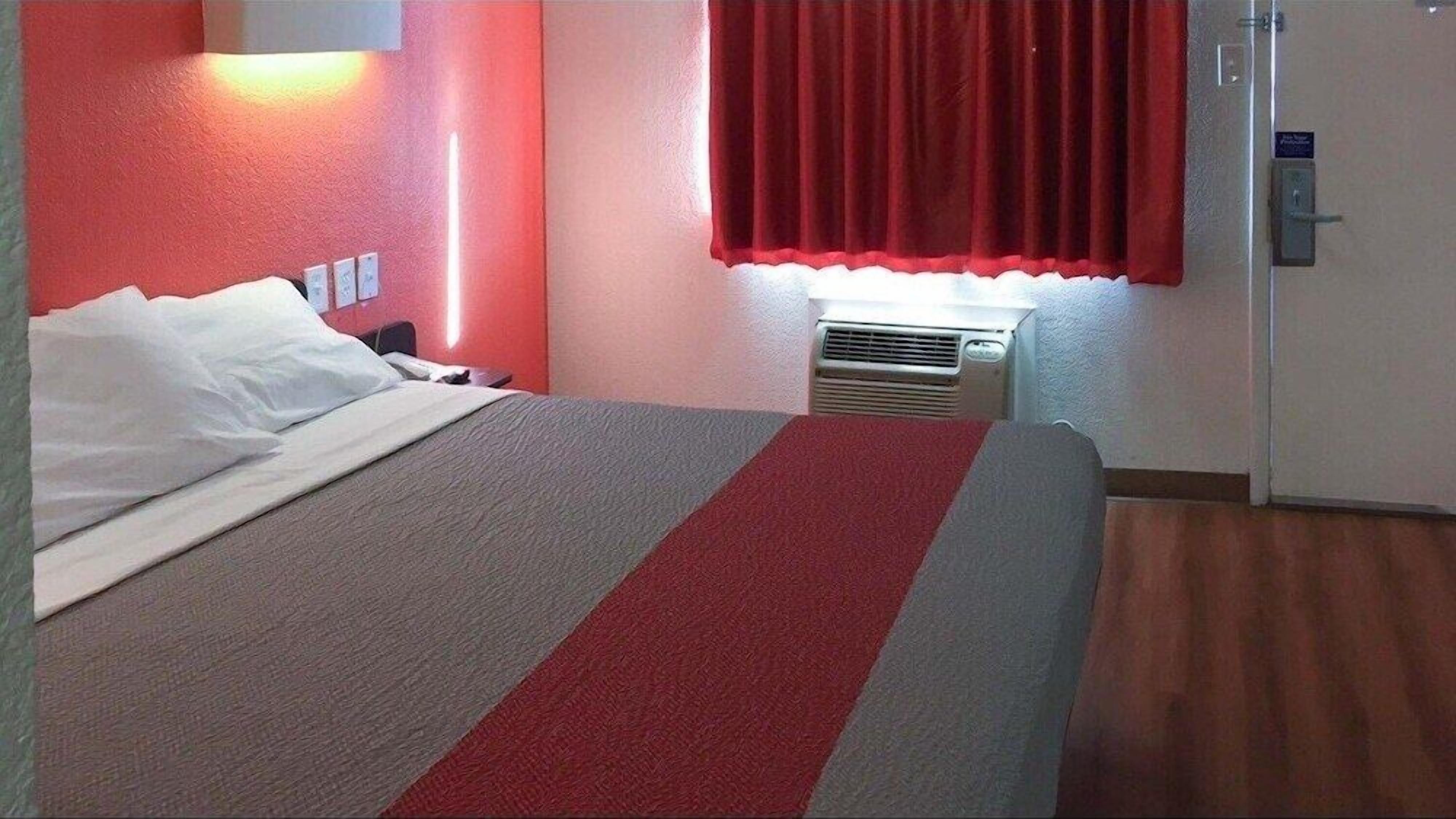 A hotel room with a bed, a red and grey striped bedspread, a white pillow, a red curtain, a wall-mounted air conditioner, and a door with a sign.