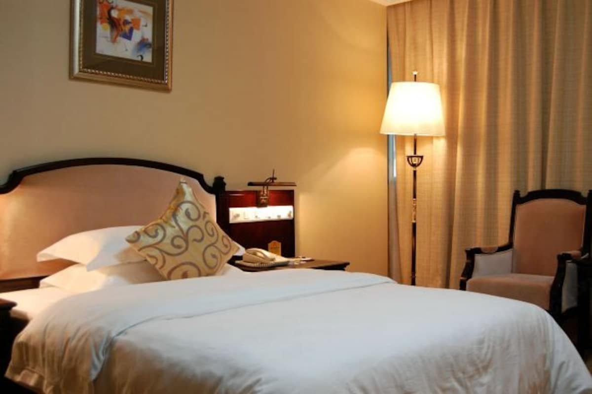 Quarto de hotel com uma cama grande, mesa de cabeceira com abajur, uma cadeira, escrivaninha com telefone e um quadro emoldurado na parede.