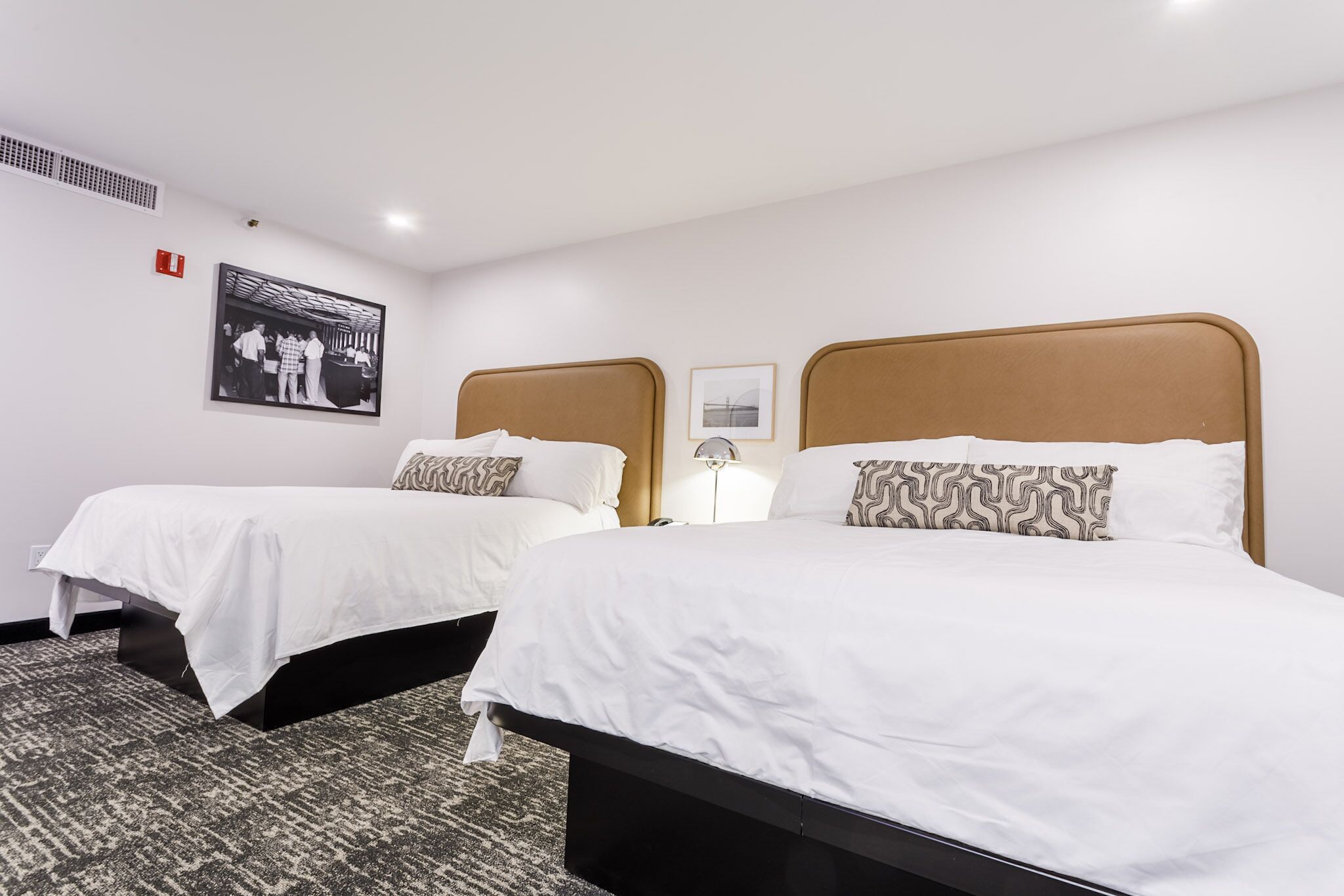 Una habitación de hotel con dos camas, un suelo con estampado, un cuadro colgado en la pared y una mesita de noche con lámpara.
