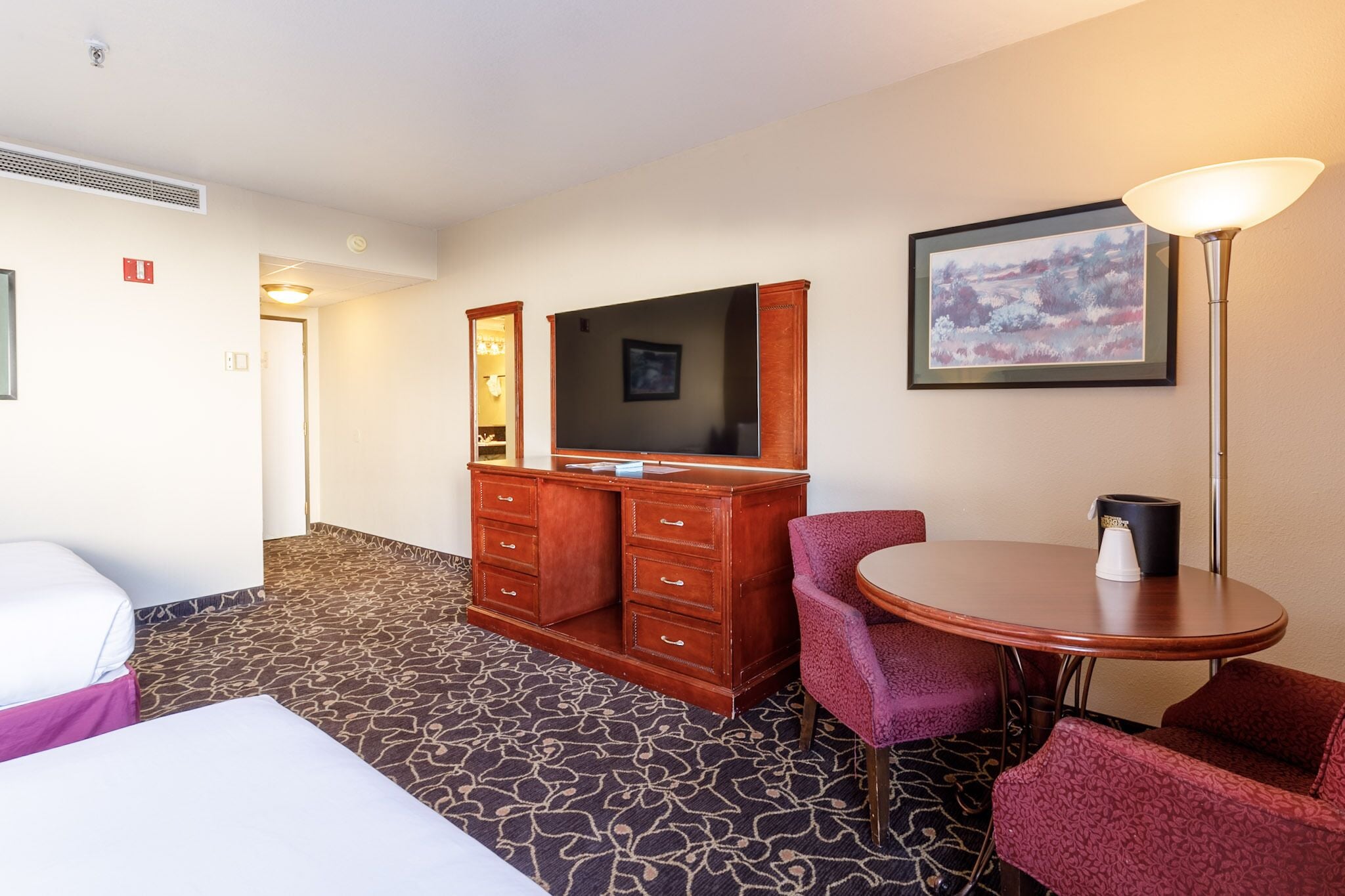 Une chambre d’hôtel avec un lit, une commode en bois, un téléviseur, un tableau encadré, une table avec une lampe et un tapis à motifs.
