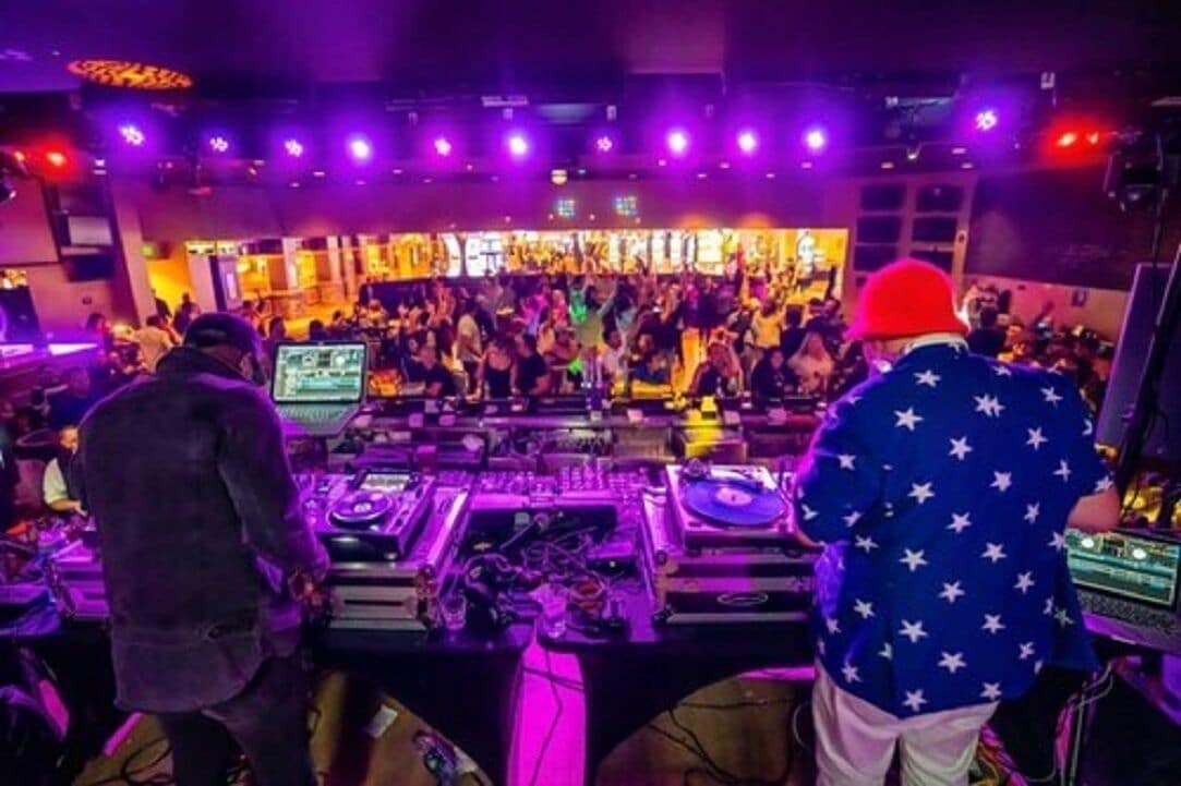 Ein DJ-Pult mit mehreren Plattenspielern und Mischpulten, umringt von einer Menschenmenge.