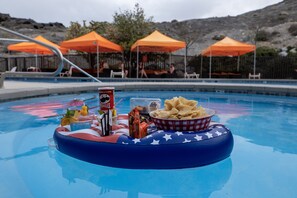 Disposición junto a la piscina con una mesa flotante que sostiene aperitivos y bebidas, rodeada de sombrillas y sillas naranjas.