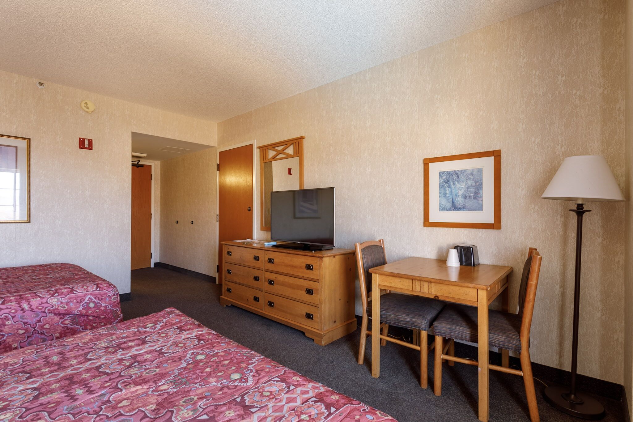 A hotel room with two beds, a wooden dresser with a television, a dining table with chairs, and a floor lamp.