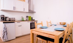 Una cucina compatta con mobili bianchi, lavastoviglie e macchina da caffè. Un tavolo da pranzo con piatti blu e bicchieri da vino è apparecchiato per due persone.