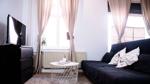 Una sala de estar moderna con un sofá negro, una mesa redonda de centro, un televisor sobre un mueble bajo y una ventana con cortinas.
