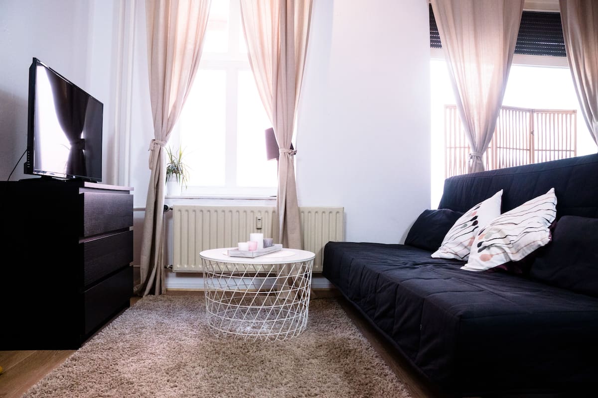 Una sala de estar moderna con un sofá negro, una mesa redonda de centro, un televisor sobre un mueble bajo y una ventana con cortinas.
