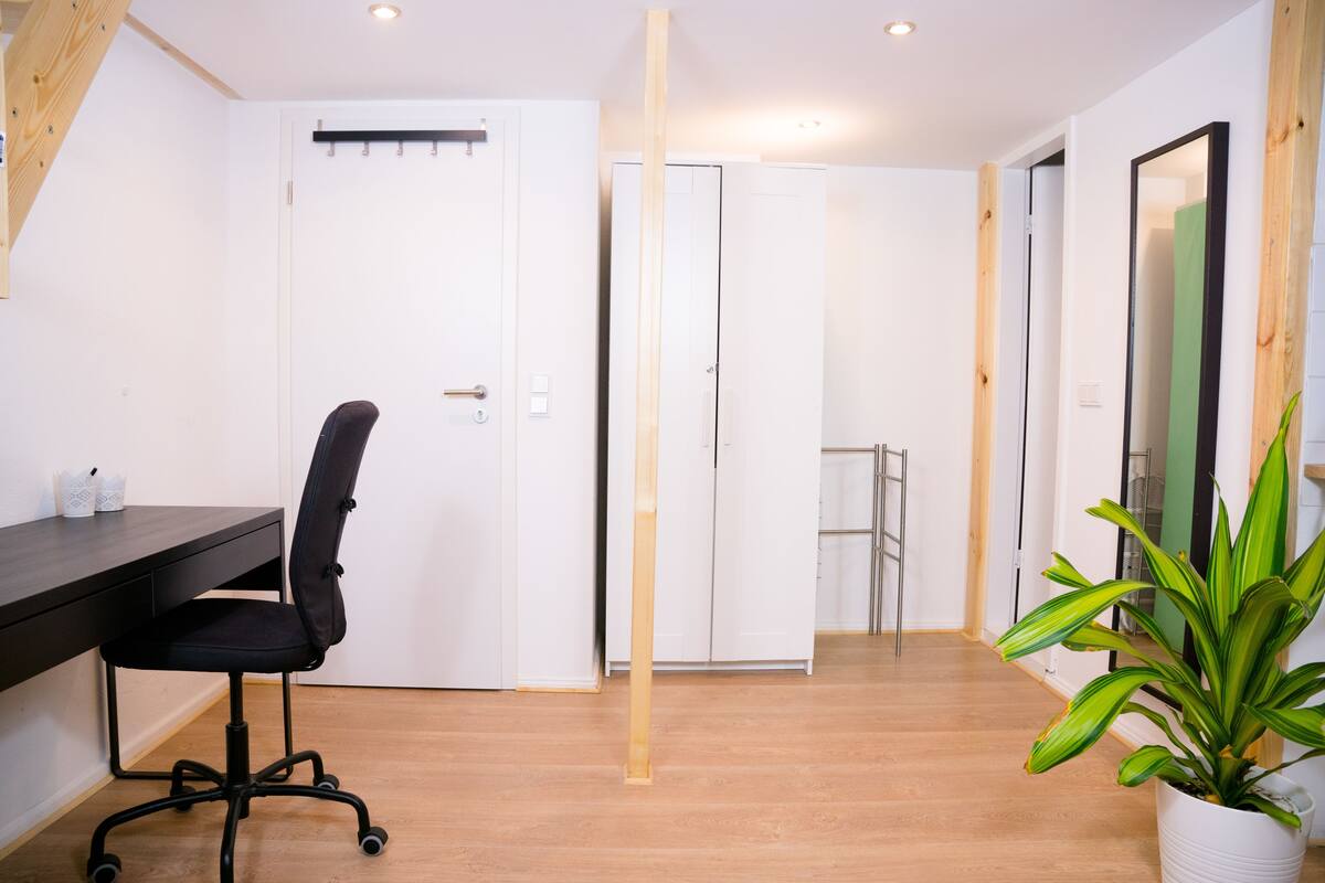 Una habitación moderna con suelo de madera, una silla de oficina negra, un escritorio blanco, una planta en maceta y un armario blanco.
