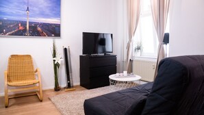Una sala moderna con un televisor de pantalla plana grande, un sofá negro, una silla de mimbre, una pequeña mesa redonda y una fotografía enmarcada de un paisaje urbano en la pared.