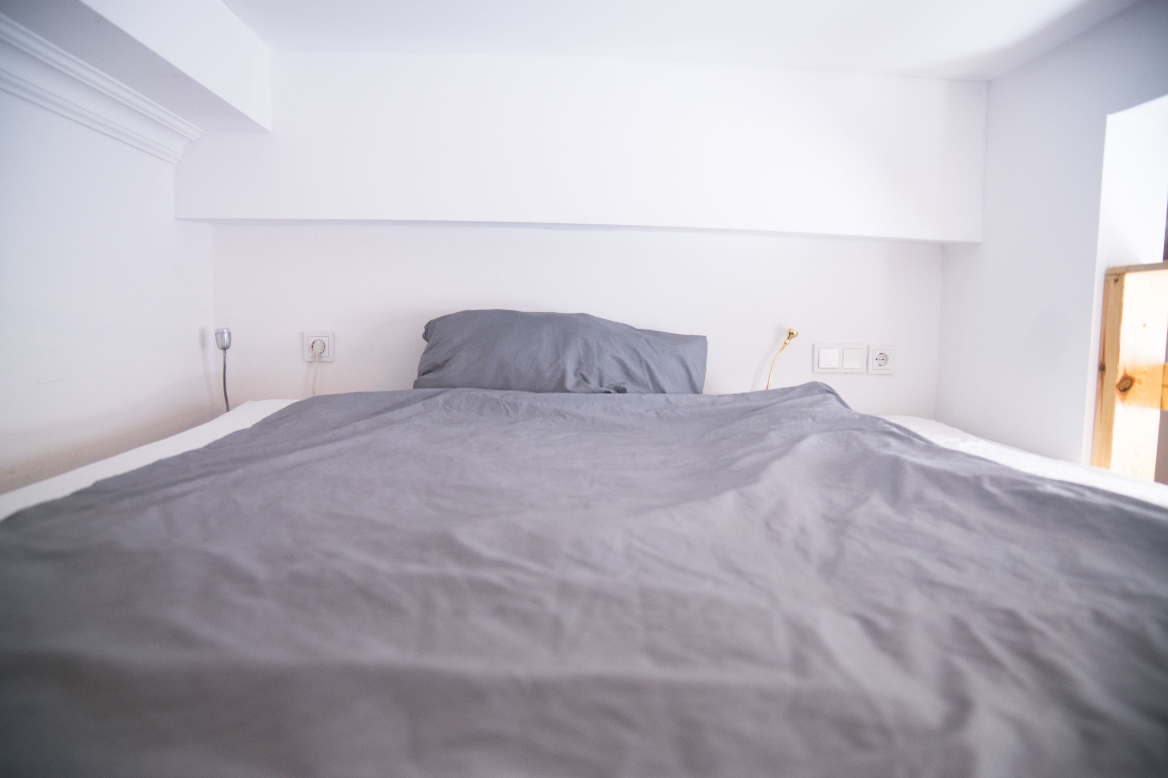 A neatly made bed with a gray blanket in a bright room with white walls and a wooden door.