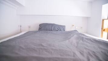 A neatly made bed with a gray blanket in a bright room with white walls and a wooden door.