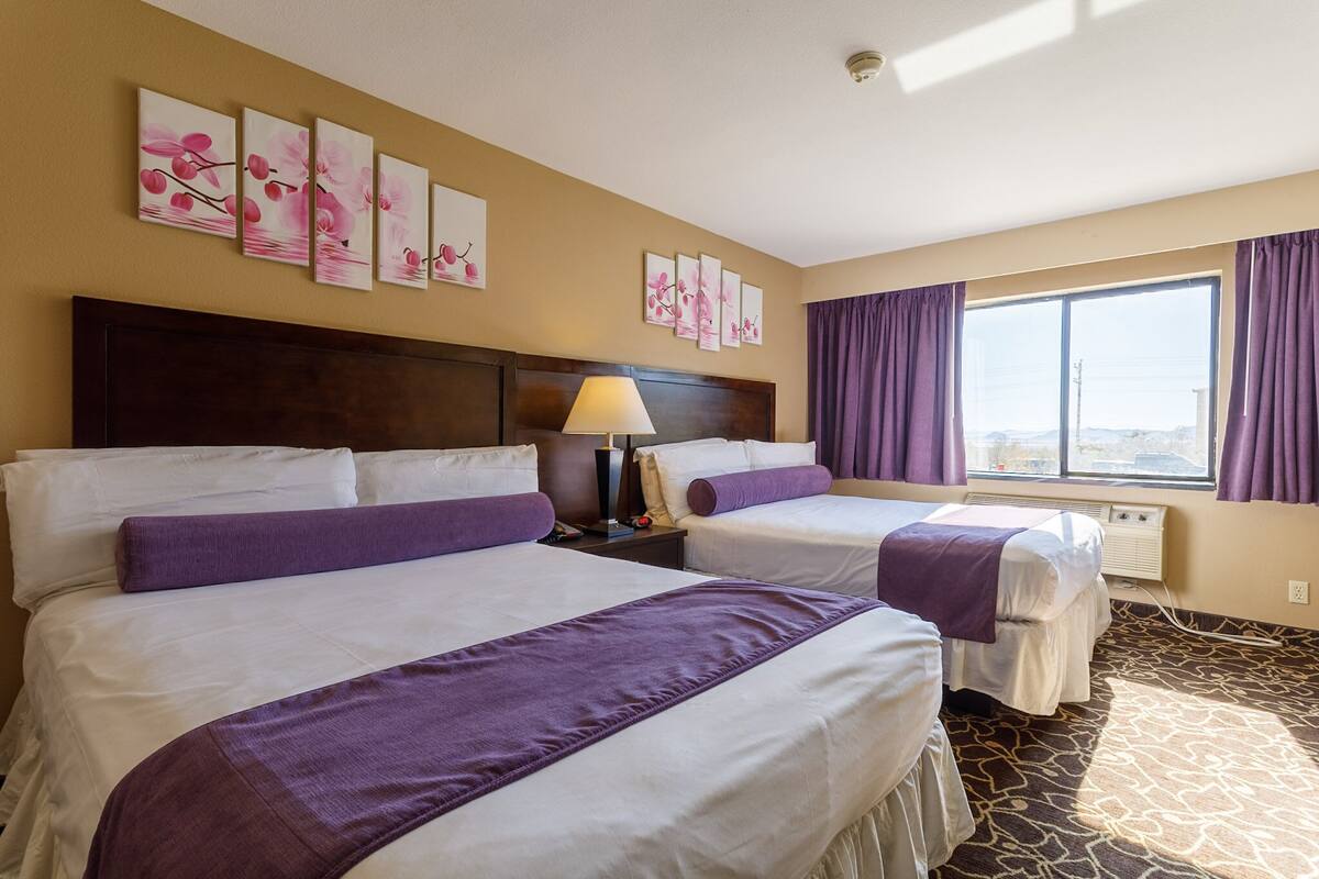 Una habitación de hotel con dos camas, un ventanal con cortinas moradas y una obra de arte floral en la pared.