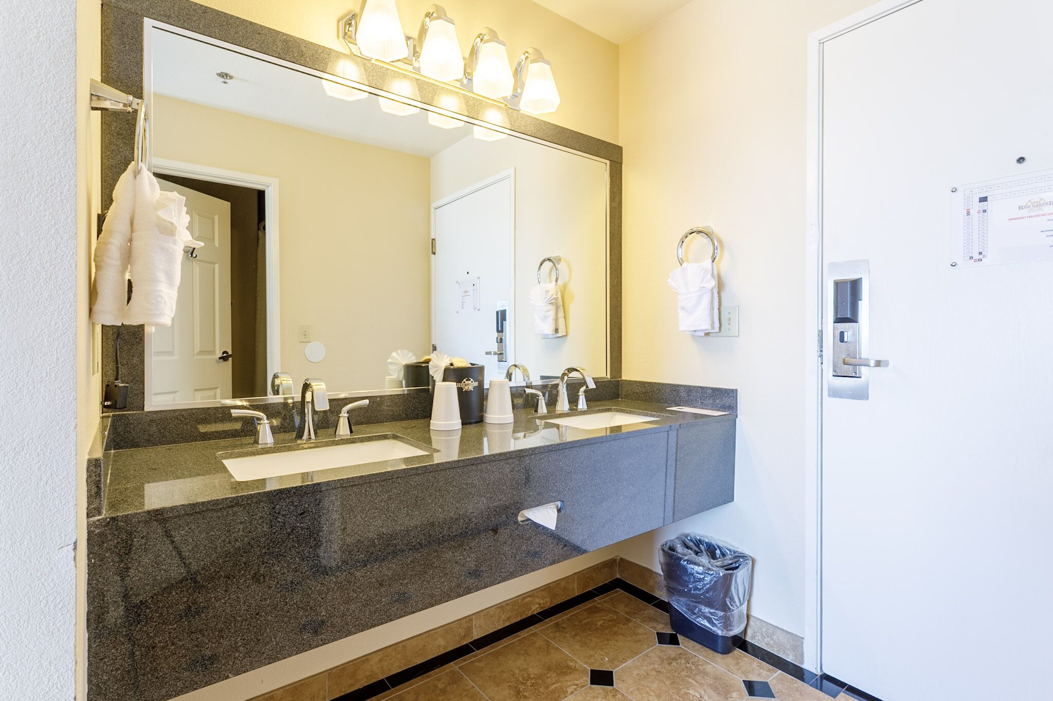 Bagno con lavabo in granito, due lavandini, uno specchio grande e un cestino.