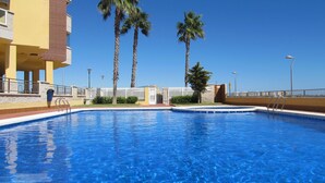 Piscina azul límpida com cerca branca, palmeiras e um edifício amarelo ao fundo.