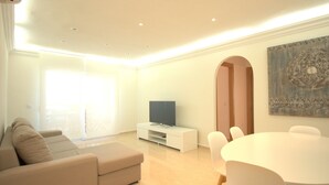 Uma sala de estar moderna com sofá, televisão em um suporte branco, mesa de jantar com cadeiras e uma grande janela que permite entrada de luz natural.