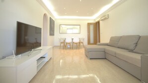 Uma sala de estar moderna com uma TV de tela plana, um sofá branco e um espaço de jantar com mesa e cadeiras.