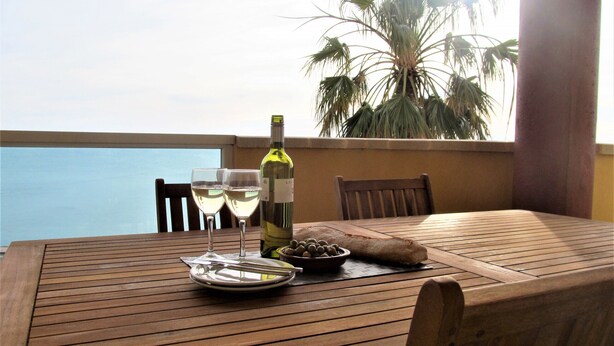 Uma mesa de madeira com duas taças de vinho, uma garrafa de vinho, um prato com azeitonas e uma tigela de frutos secos numa varanda com vista para o mar.