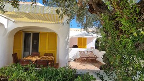 A covered outdoor seating area with a table and chairs, surrounded by greenery and a white building with yellow shutters. - Aldeia do Golf- Family with Pool and Wi-fi (Vilamoura)