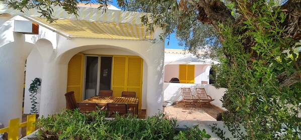 A covered outdoor seating area with a table and chairs, surrounded by greenery and a white building with yellow shutters.