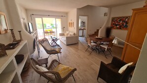 A spacious living area with a sofa, dining table, wicker chairs, and a kitchen area visible through an open door. - Aldeia do Golf- Family with Pool and Wi-fi (Vilamoura)